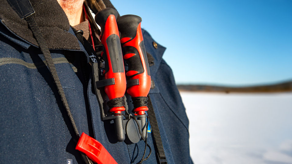 Isdubbar hänger runt halsen på en person. I bakgrunden syns en frusen sjö och blå himmel. Foto. 