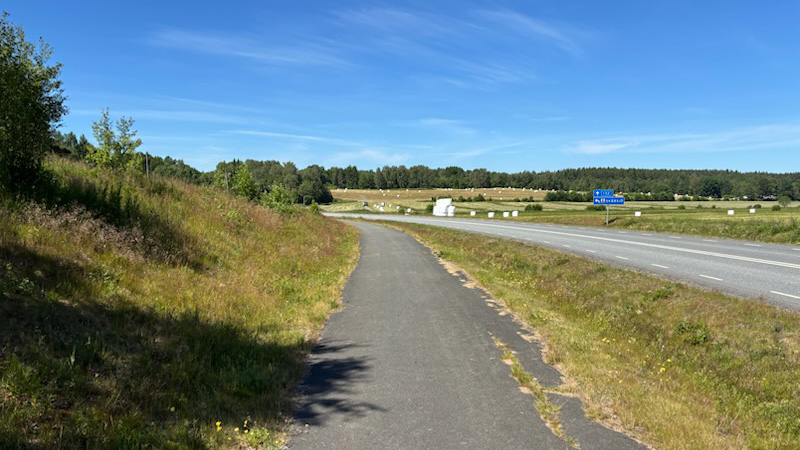 En asfalterad gång- och cykelväg löper längs en landsväg under en klarblå himmel. I bakgrunden syns gröna fält, skog och vita höbalar utspridda på ängen. Foto