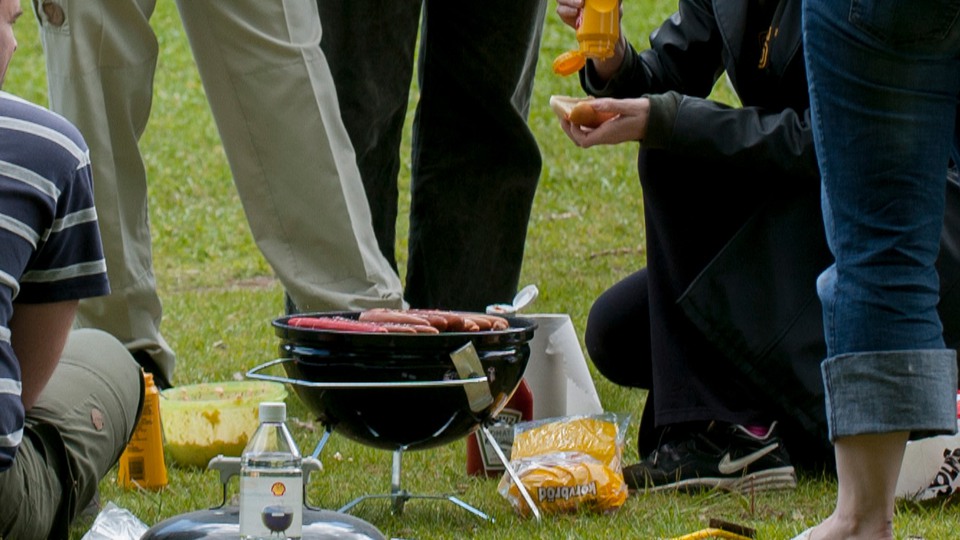Personer som står runt el kolgrill, en person lägger senap på en korv. Foto 