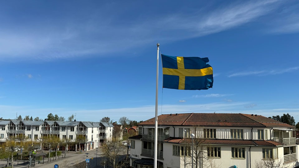 Svensk flagga är hissad och vajar i vinden. I bakgrunden syns byggnader och hustak. Foto.
