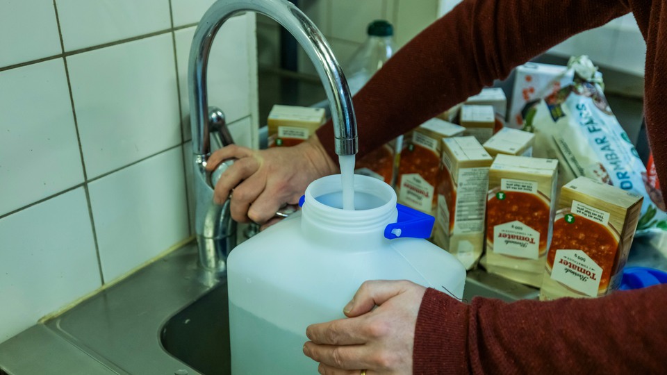 Person fyller en vattendunk med vatten. I bakgrunden syns tomma förpackningar med konserver. Foto. 