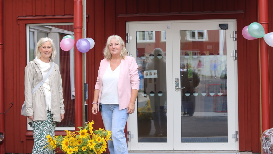 Två kvinnor som står framför en röd byggnad med gula blommor. Foto 
