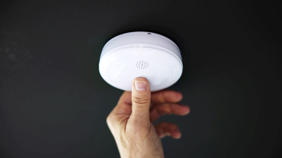 Person trycker på testknappen på en brandvarnare som hänger i taket. Foto. 
