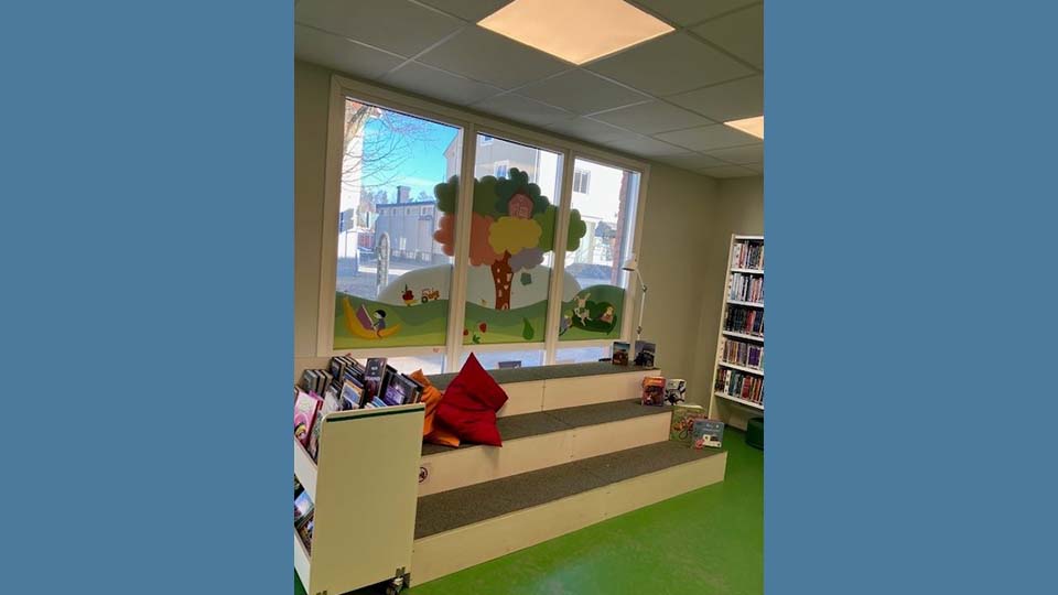 Blå bakgrund. Bild av läshörna på biblioteket, en tygklädd låg trappa framför ett stort fönster och grönt golv.