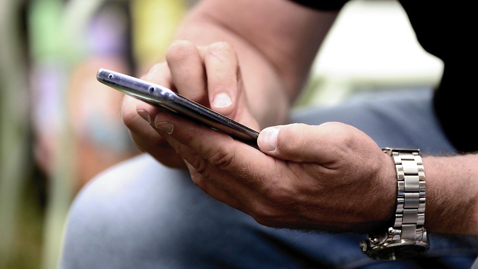 ärbild på en person som håller i en smartphone med båda händer och använder pekskärmen. Personen bär en metallklocka på handleden, och bakgrunden är suddig. Foto 