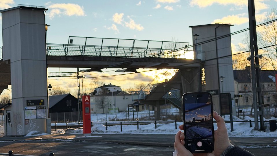 En gångbro över järnväg i vintermiljö vid solnedgång. I förgrunden håller en hand en mobil som fotograferar bron, snö på marken och byggnader i bakgrunden. Foto