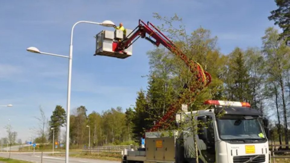 En servicearbetare står i en skylift monterad på en lastbil och utför underhåll på en gatlampa längs en väg. Bakom syns träd och en blå himmel. Foto