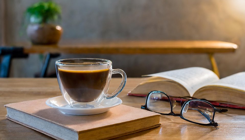 På ett bord står det en genomskinlig kaffekopp. Bakom ser men lite av en bok och på boken är det ett par glasögon. Fokuset ska vara på kaffekoppen