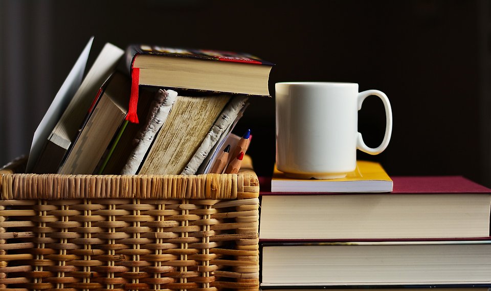 Böcker staplade på hög i en korg. Kaffekopp står placerad på högen med böcker. 