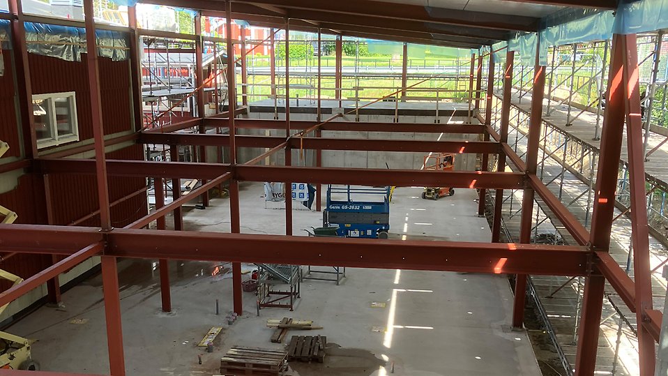Pågående byggprojekt med stålkonstruktion och ställningar. Betonggolvet är färdiggjutet och byggställningar finns längs sidorna. Byggmaskiner och material är placerade inne i strukturen. Foto