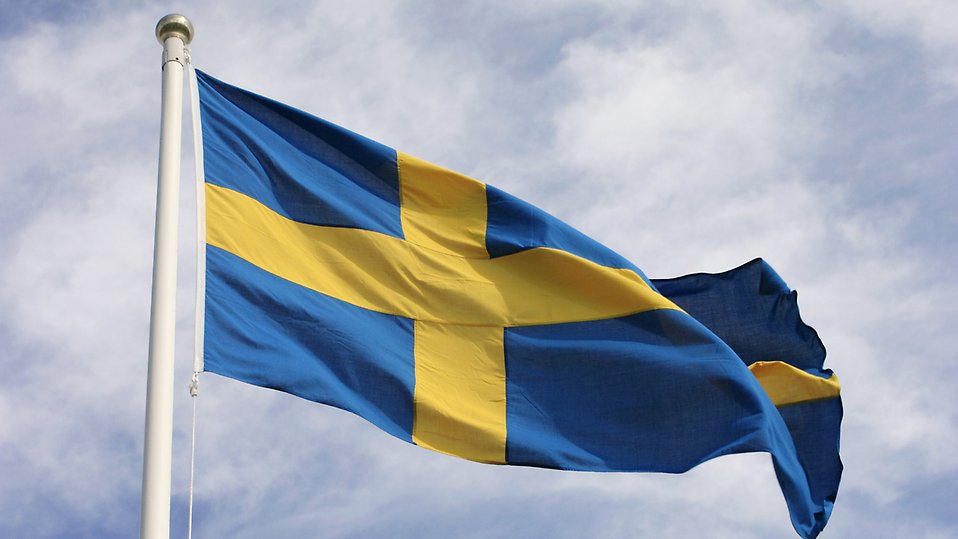 Svensk flagga på en flaggstång blåser i vinden. Foto.
