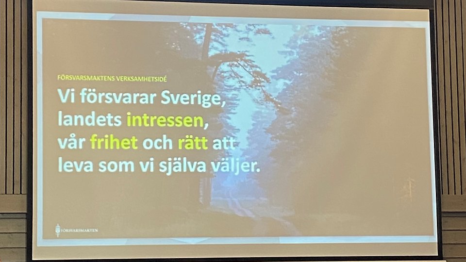 En projektorduk visar en bild med text om Försvarsmaktens verksamhet mot en bakgrund av en skogsstig. Foto 