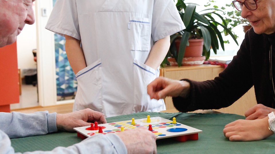 Två personer spelar ett brädspel med färgade pjäser vid ett bord, medan en person i vårdkläder står bredvid. På bordet finns en grön duk och i bakgrunden syns krukväxter. Foto