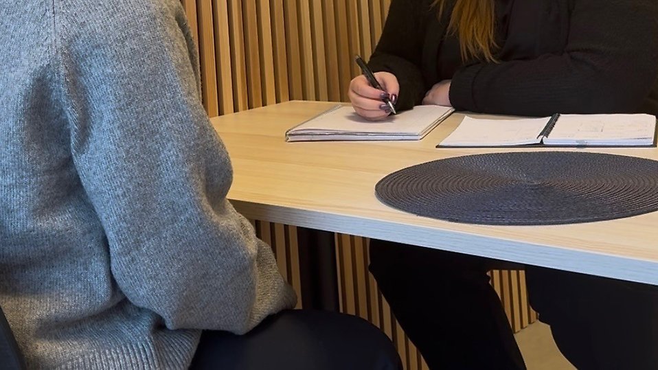 Två personer sitter vid ett ljust träbord i samtal. Den ena personen, i grå tröja, sitter med ryggen mot kameran medan den andra, klädd i svart, antecknar i ett block. På bordet ligger ett runt mörkgrått bordstablett. Bakgrunden består av en vägg med vertikala träpaneler. Foto 