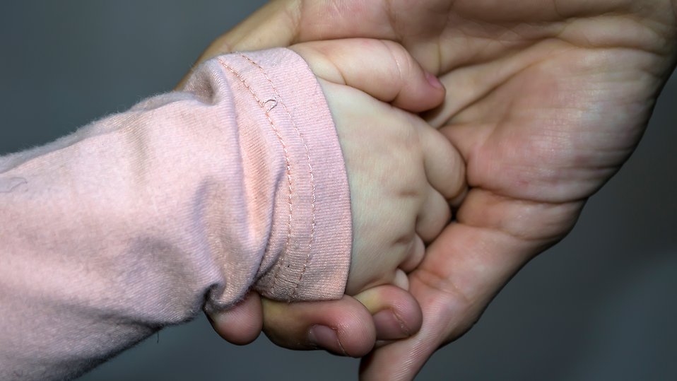 En vuxen hand håller en barns hand vars tröja är ljusrosa. Foto